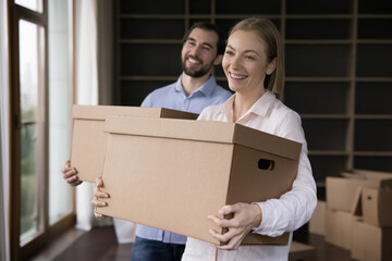 Young wife and husband holding boxes with personal belongings pose in unfurnished living room. Happy homeowner family portrait, moving day, start new life at own or rented dwelling, repairs concept