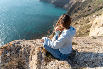 Naklejka premium Woman tourist enjoying the sunset over the sea mountain landscape. Sits outdoors on a rock above the sea. She is wearing jeans and a blue hoodie. Healthy lifestyle, harmony and meditation