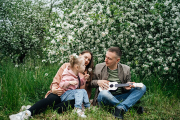  family mom mom baby daughter in the garden blooming apple trees, father playing the ukulele