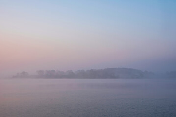 Fototapeta premium Beautiful landcape image of Spring sunrise over reservoir lake with dawn glow spreading aross the water with low mist adding atmosphere