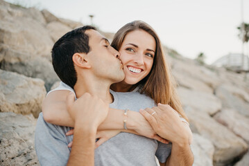 Fototapeta premium A close photo of a smiling woman who is hugging her Hispanic boyfriend at sunset in Valencia. A man is kissing his wife, sitting on the stones of a breakwater in Spain in the evening.