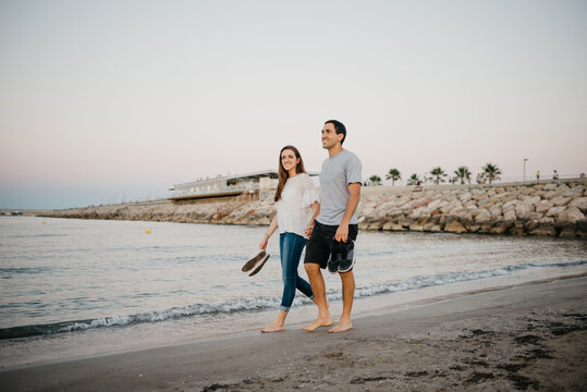 A full-length photo of a Hispanic man with his happy girlfriend who are strolling barefoot on a sandy beach near the Balearic sea in Spain in the evening. A couple of tourists on a date in Valencia