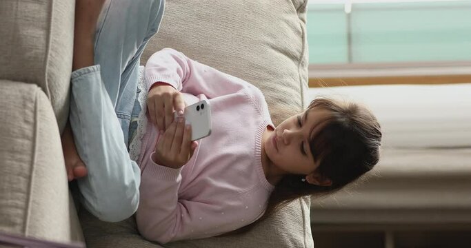 Young gen Z use modern wireless technology, gadgets overuse, bad life habit, lifestyle concept. Vertical view little girl sits on sofa holds smart phone, chat online, share text messages via messenger