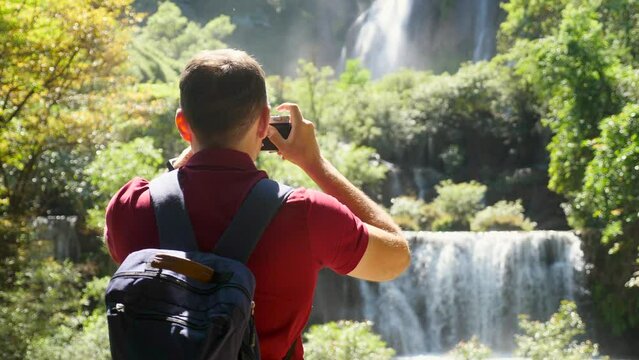 Happy male tourist in trip, journey. Young man blogger, zoomer traveler make photo for social media of amazing waterfall, nature on trendy retro camera. Wanderlust, travel concept. Cinematic shot.