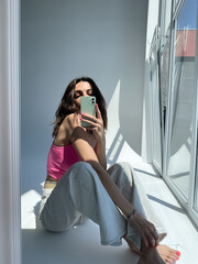 Fit tanned woman in jeans and pink crop top, summer street wear, take photo selfie on phone in mirror for social media, stories, vertical.