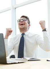 Happy young businessman raising hands in front of laptop at office desk