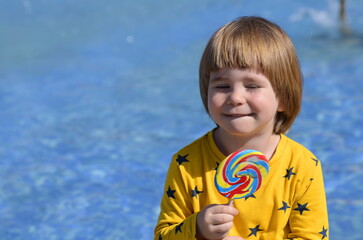 A little boy eats a big colorful lollipop while sitting by the pool. Concept: bright summer, family vacation, travel with children, resort, water park. The child rejoices in the summer, smiles 