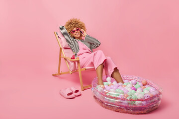 Young positive woman with curly hair poses on comfortable deck chair makes phone call wears sunglasses striped jumper and overalls dreams about summer vacation isolated over pink background.