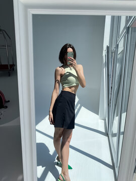Fit tanned woman in classic short black skirt, light green crop top and heels, summer fashion stylish wear, take photo selfie on phone in mirror for social media, stories, vertical.
