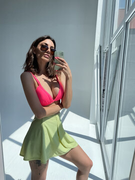 Fit tanned woman in light green beach skirt, sunglasses, bright pink swimsuit bra, summer fashion stylish wear, take photo selfie on phone in mirror for social media, stories, vertical.