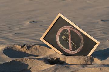 Pizarra negra con marco de madera de color claro con el símbolo de prohibido fumar escrito con tizas de colores, sobre la arena de una playa  durante el atardecer un día de verano - obrazy, fototapety, plakaty