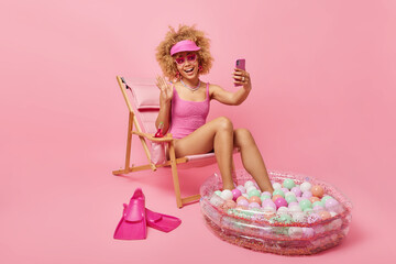 Positive carefree woman dressed in swimsuit during summer time makes online call via smartphone waves hello in smartphone camera sits on deck chair against pink background. Good rest concept
