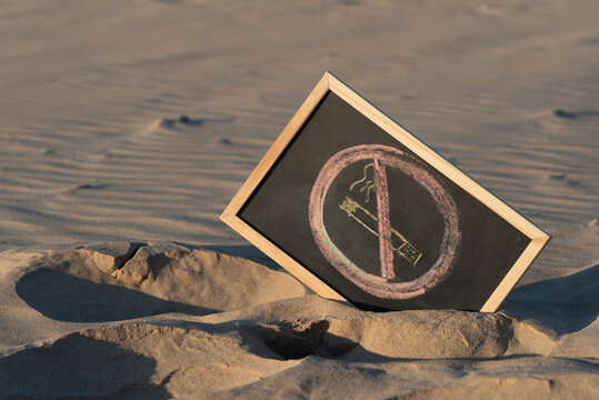 Pizarra negra con marco de madera de color claro con el s&iacute;mbolo de prohibido fumar escrito con tizas de colores, sobre la arena de una playa  durante el atardecer un d&iacute;a de verano