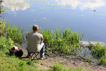 Fisherman sitting near the water with a fishing rod, rear view. Man angling on the lake coast at...