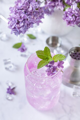 Lilac drink, infused drink from lilac flowers. Organic natural daikiri cocktail or mocktail, lilac lemonade with ice on a marble table.