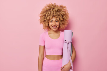 Cheerful woman with curly hair wears sportive outfit holds rolled mat ready for fitness training at...