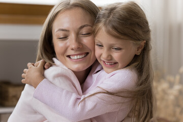 Loving little 6s daughter cuddles her young mum, close up view beautiful faces, enjoy embraces and moment of tenderness. Kid congratulates mom with Happy Mother Day, family bond and ties, love concept
