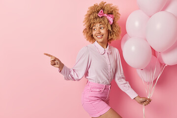 Obraz na płótnie Canvas Cheerful woman with curly hair wears blouse and shorts holds bunch of inflated balloons points away into distance prepares for party isolated over pink background empty space for your promotion