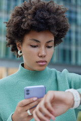 Fototapeta na wymiar Vertical shot of serious curly haired young woman checks time on smartwatch uses mobile phone wears casual jumper poses outdoors against blurred background. People technology lifestyle concept