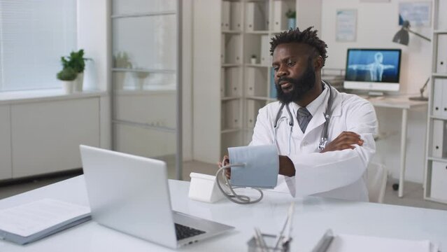 Modern young adult African American doctor wearing white coat sitting at desk working with patient remotely