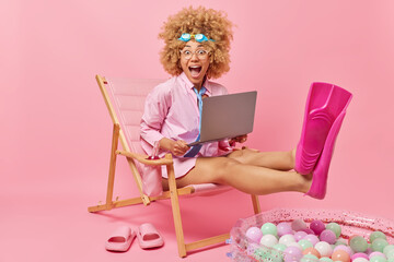 Joyful surprised curly woman dreams about summer vacation wears goggles shirt and flippers works on laptop computer poses on deck chair against pink background. People holidays tourism concept