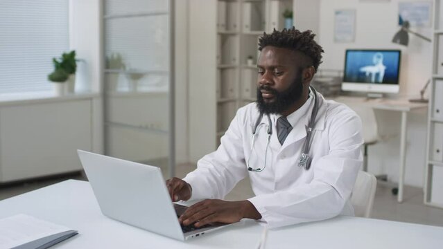 Modern young adult African American doctor wearing white coat sitting at desk typing something on laptop then looking at camera