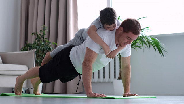 happy father doing push up exercise with son on back at gym. little boy hugs dad from behind