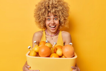 Positive surprised woman carries portable fridge with bottles of cool beverage and citrus fruits going to spend all summer day at beach wears casual t shirt isolated over vivid yellow background