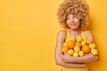 Indoor shot of pleased curly haired young woman embraces big heap of oranges and lemons full of...