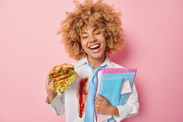 Positive female student eats delicious hamburger carries folders with sticker has tasty snack...