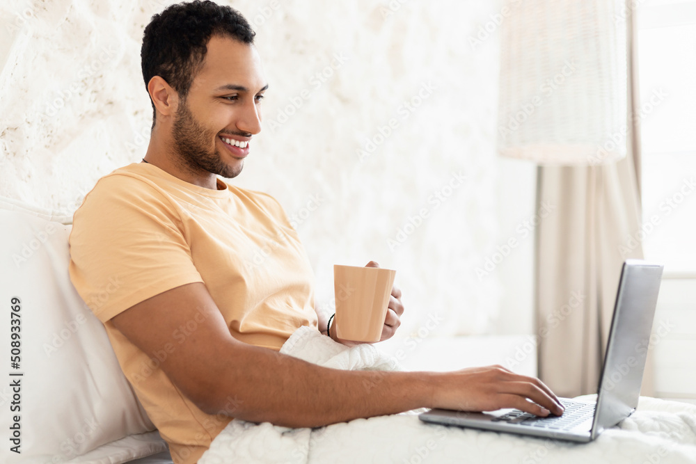 Wall mural Arabic Guy Using Laptop Drinking Coffee In Bed At Home