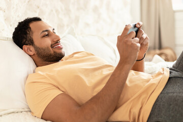 Obraz na płótnie Canvas Cheerful Arabic Guy Using Cellphone Playing Game Lying At Home