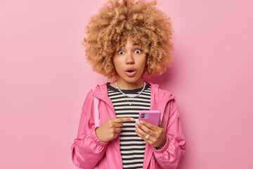 Shocked impressed woman with curly hair points at smartphone screen reads surprising news on webpage indicates at digital cellular dressed in jacket isolated over pink background. Omg look at this