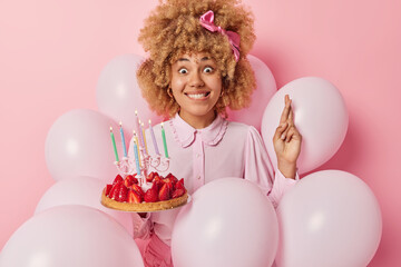 Fototapeta na wymiar People holidays and celebration concept. Cute curly haired woman dressed in festive clothes holds delicious strawberry cake with burning candles keeps fingers crossed poses around inflated balloons