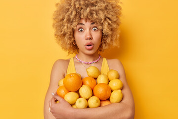 Impressed shocked woman with curly blonde hair embraces heap of juicy fresh oranges and lemons keeps mouth opened from wonder isolated over vivid yellow background. Citrus fruits full of vitamins - obrazy, fototapety, plakaty