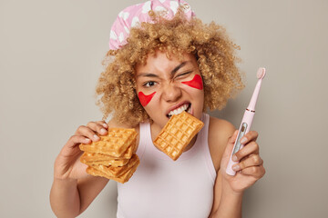 Photo of curly haired woman eats delicious homemade waffle holds electric brush for cleaning teeth applies red beauty patches to remove wrinkles dressed in casual t shirt isolated on grey studio wall - obrazy, fototapety, plakaty