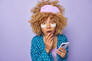 Stunned curly haired woman stares bugged eyes cannot believe in shocking news applies beauty patches under eyes dressed in pajama sleepmask holds mobile phone isolated over purple studio wall