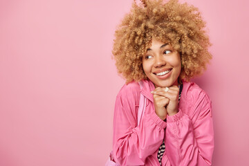 Pretty thoughtful woman has glad expression keeps hands under chin smiles toothily dressed in jacket focused away isolated over pink background with blank space for your promotional content.