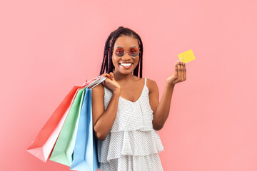 Cheery young black lady in dress and sunglasses holding credit card and shopping bags on pink studio background