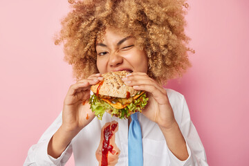 Tasty unhealthy meal. Hungry curly haired young woman bites delicious hamburger break diet wears...