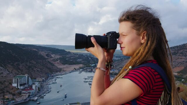 Attractive girl with dreadlocks takes pictures of river in gorge with ships and buildings on shore. Profile side view. Young woman photographer shoot photos of water landscape with mountains