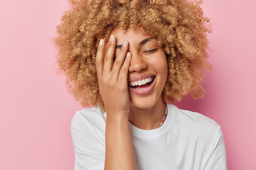 Fototapeta Headshot of positive carefree woman makes face palm smiles toothily keeps eyes closed has pleasant feelings dressed in white t shirt isolated over pink background. People and emotions concept obraz