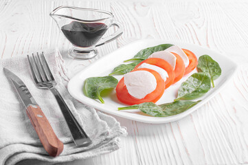 caprese salad made from pieces of tomato, mozzarella and spinach leaves lies in a white plate next to a fork and knife and balsamic sauce on a white wooden background