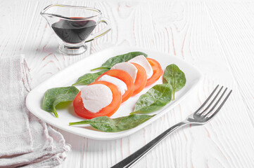 caprese salad made from pieces of tomato, mozzarella and spinach leaves lies in a white plate next to a fork and balsamic sauce on a white wooden background