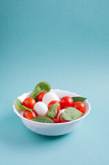 caprese salad of cherry tomatoes, mozzarella balls and spinach leaves lies in a white plate on a blue background with copy space