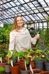 Portrait of a young woman in a greenhouse. She looking at camera, smiles and holds a water sprinkler in her hands.