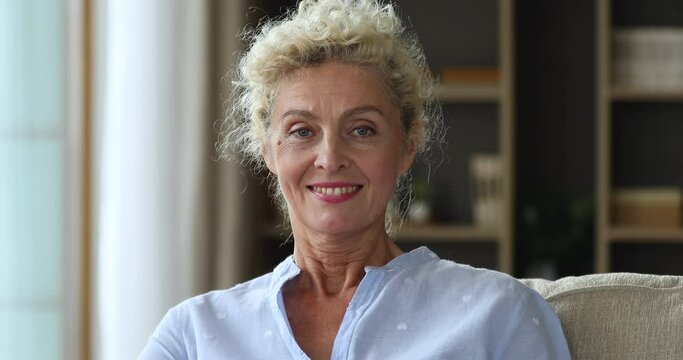 Head shot close up beautiful middle-aged woman smile staring at camera relax on sofa in living room. Good-looking baby boomer generation female portrait, carefree retirement, happy homeowner concept