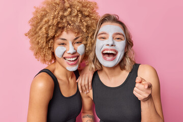 Indoor shot of happy female friends undergo beauty procedures for skin treatment apply clay masks laugh positively dressed in casual black t shirt point at camera isolated over pink studio background
