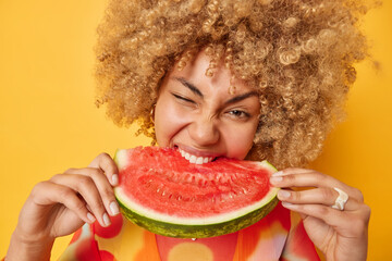 Close up shot of curly haired cheerful woman holds bigs slice of juicy watermelon enjoys eating...
