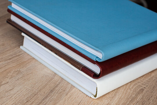 Three stylish photo books with leather covers, white, burgundy and blue, of different thicknesses, lie on a brown wooden tabletop, indoors.